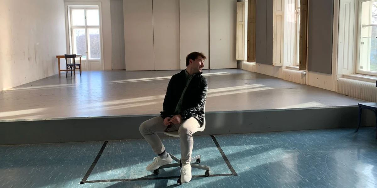 Simon Dworaczek sitzt vor einer leeren Theaterbühne im natürlich beleuchteten Probenraum.